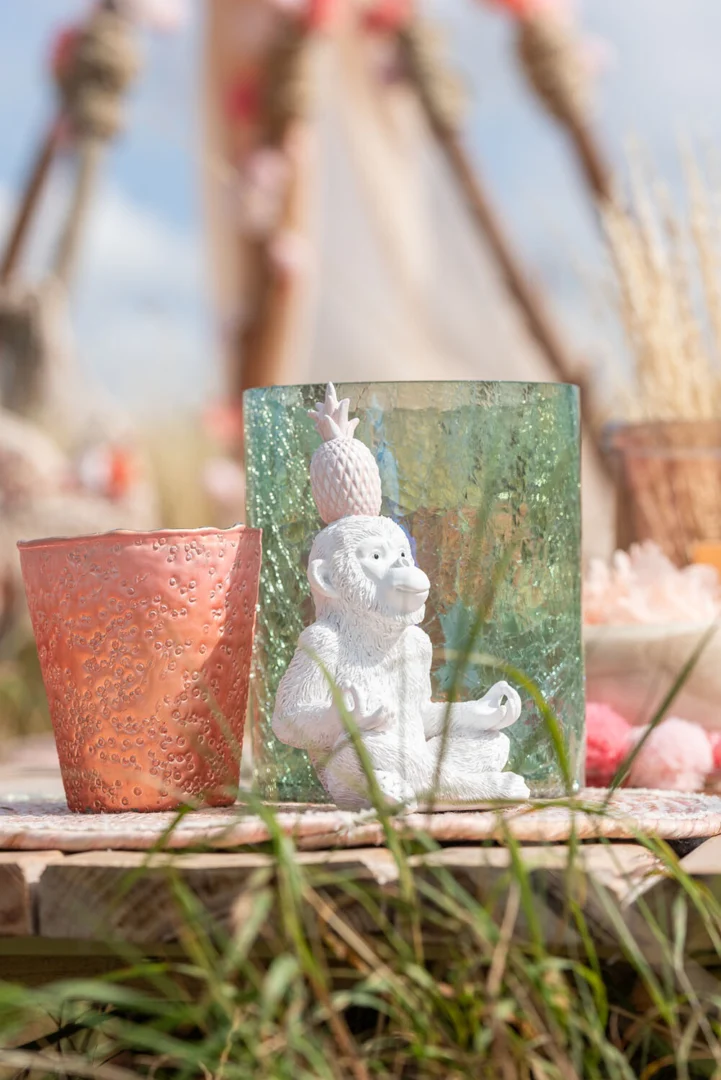 Een wit apenbeeldje met een ananas op zijn hoofd staat naast een kandelaar met roze structuur en de Kaarsenhouder Orkaan Crackle Glas Groen Groot op een houten buitenoppervlak met gras op de onscherpe achtergrond.