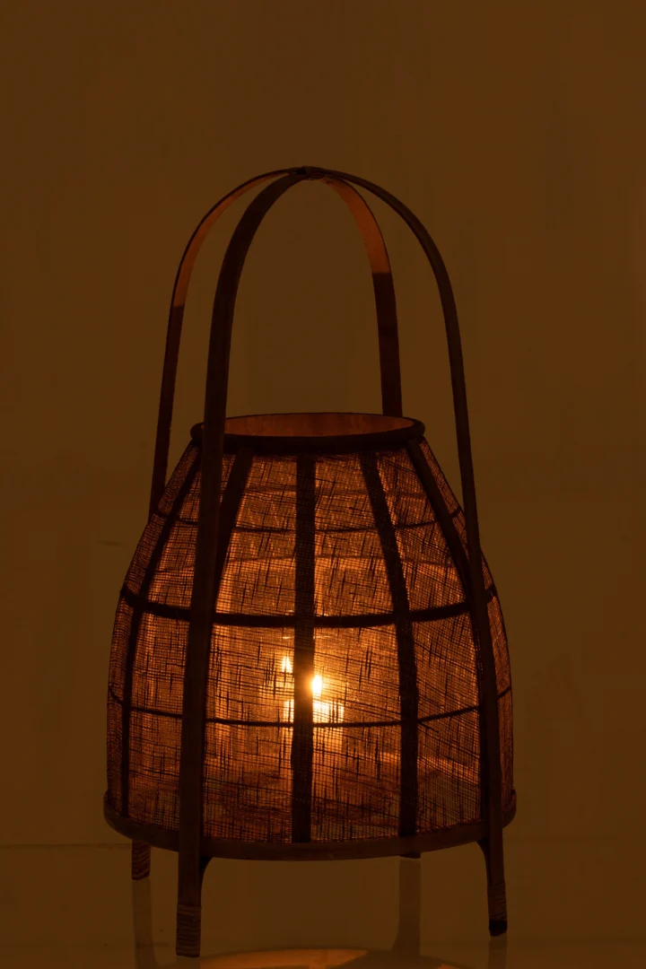 De Lantaarnkap Bamboe Natuurlijk Medium heeft een gloeilamp binnenin, die een warme, zachte amberkleurige gloed uitstraalt door het natuurlijke bamboevlechtwerk tegen een schemerige achtergrond.