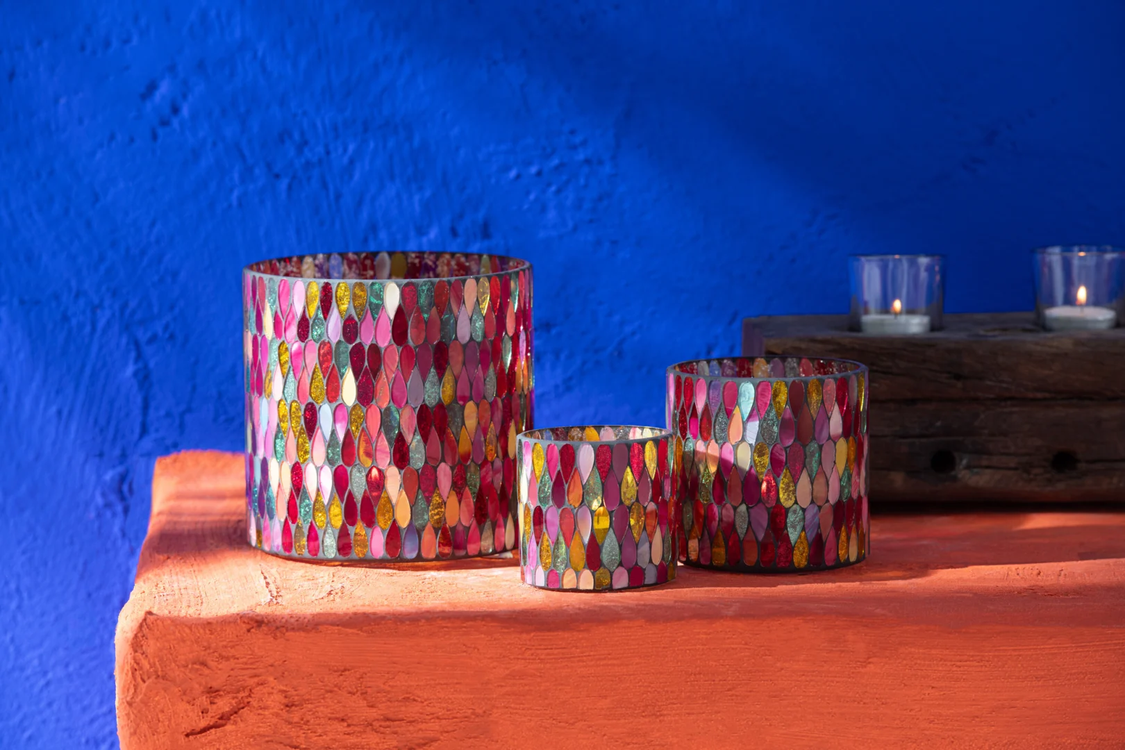 Drie Kaarsenhouder Hurricane Mozaïek Glas Mix Medium kandelaars met levendige mozaïekpatronen staan op een oranje oppervlak voor een blauwe muur. Daarachter staan twee kleine glazen waxinelichthouders op een houten dienblad.