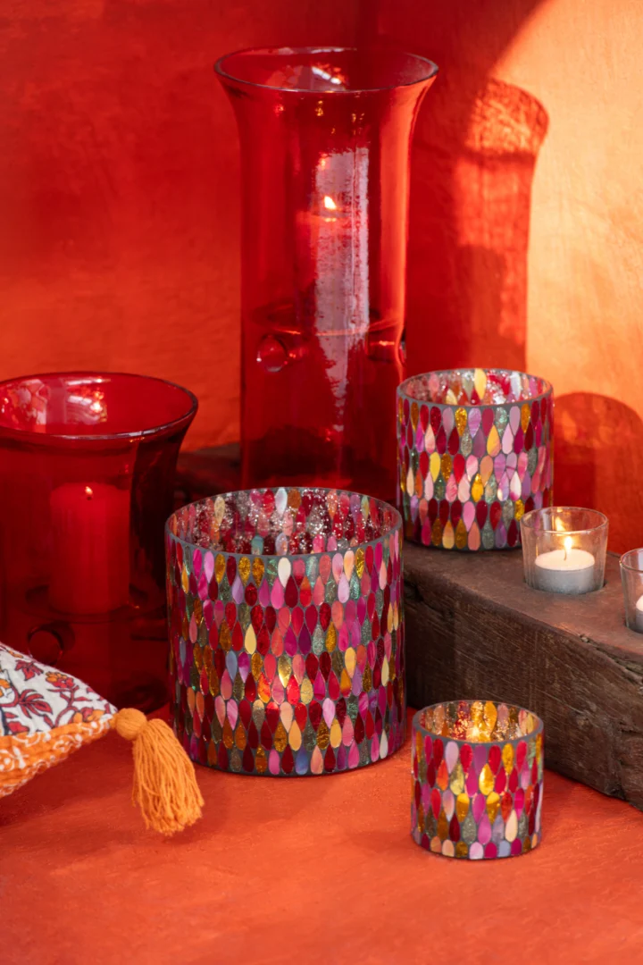 De Kaarsenhouder Hurricane Mozaïek Glas Mix Medium, met kleurrijke mozaïek- en massief rode glasontwerpen, zijn gerangschikt met brandende waxinelichtjes op een houten oppervlak in een warme, oranjekleurige omgeving.