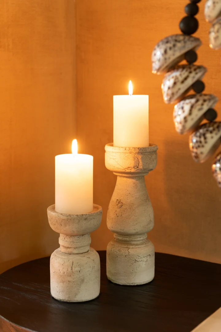 Twee witte pilaarkaarsen branden op Kaarsenhouder Terracotta Beige Groot, rustieke crèmekleurige stenen houders, op een donker oppervlak. De warm verlichte achtergrond toont een deel van een decoratief kralenobject rechts.