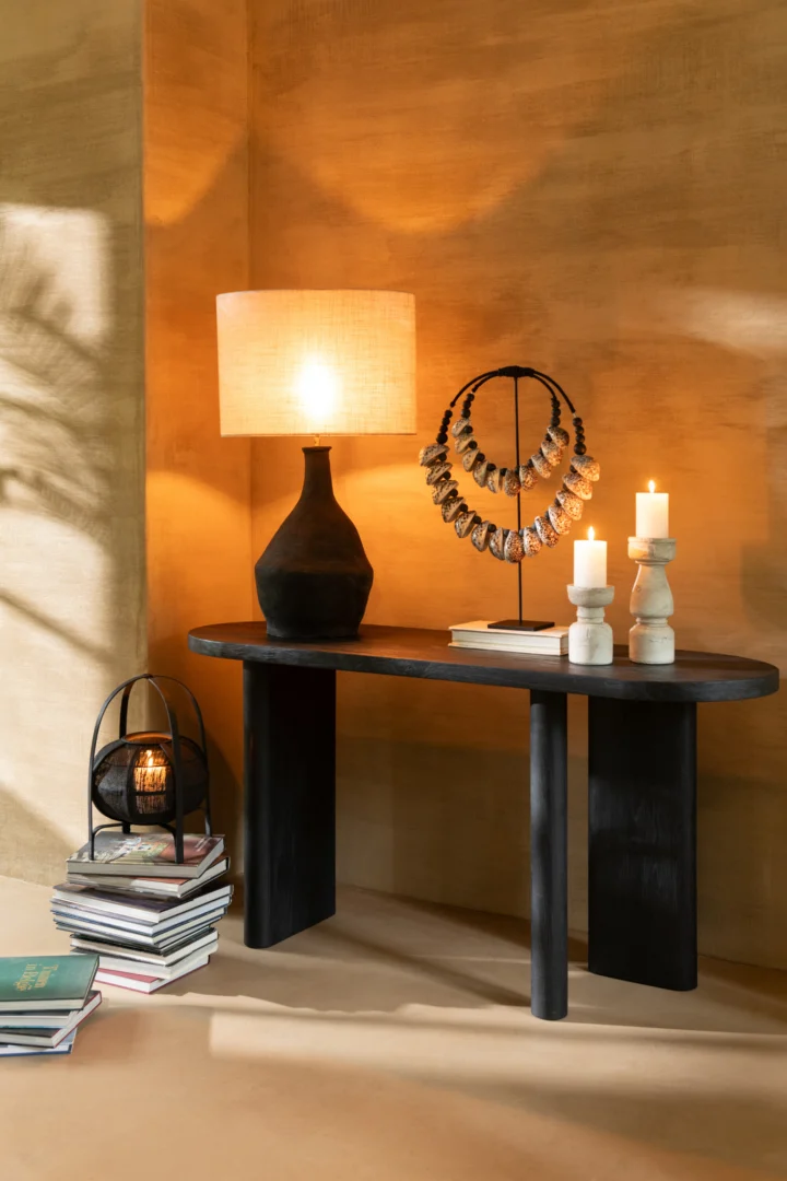 Een zwarte consoletafel staat tegen een beige muur met warme verlichting, met de Kaarsenhouder Terracotta Beige Groot met kaarsen, een donkere keramische lamp, een display met kettingen en stapels boeken.