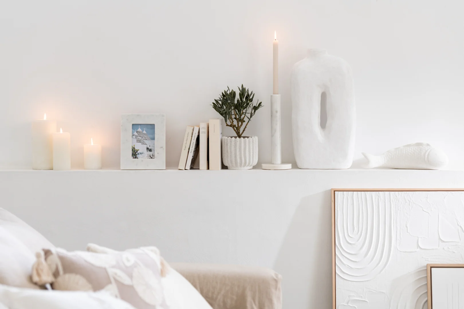 Een minimalistische witte plank toont de Kaarsenhouder Marmer Wit Set 3 met brandende kaarsen, een kleine potplant, boeken, een ingelijste foto en een sculpturale witte vaas. Een kunstwerk met textuur en licht beddengoed zijn zichtbaar op de voorgrond.