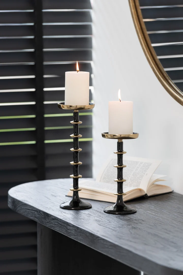 Twee witte pilaarkaarsen staan op Kaarsenhouder Bal Metaal Zwart Goud Klein kandelaars op een donkere houten tafel naast een open boek, met zwarte raamgordijnen en een deel van een ronde spiegel op de achtergrond.