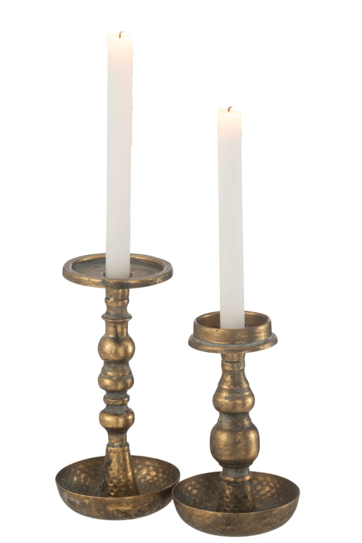 Twee hoge witte kaarsen staan in Kaarsenhouder Jere Metaal Goud kandelaars - antieke messing houders met brede, ronde voet en een afwerking met structuur. De ene houder is iets hoger dan de andere.