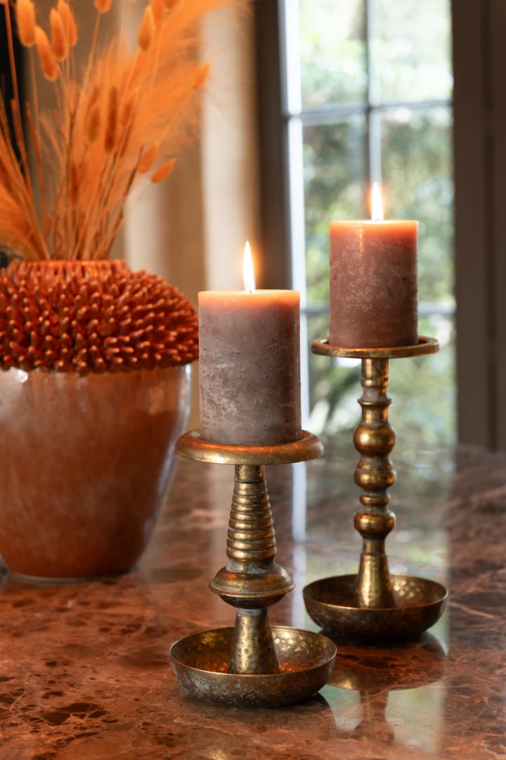 Twee brandende bruine pilaarkaarsen op Kaarsenhouder Jere Metaal Goud houders staan op marmer. Op de achtergrond staat een vaas met gedroogde oranje en beige grassen bij een raam met groen buiten.
