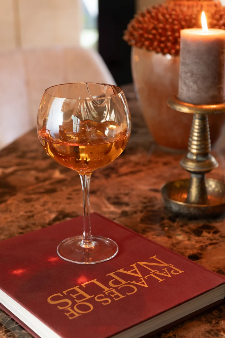 Een glas amberkleurige drank met ijs rust op een boek op een marmeren tafel, naast de Kaarsenhouder Jere Metaal Goud kandelaar en een vaas met gedroogde bloemen.