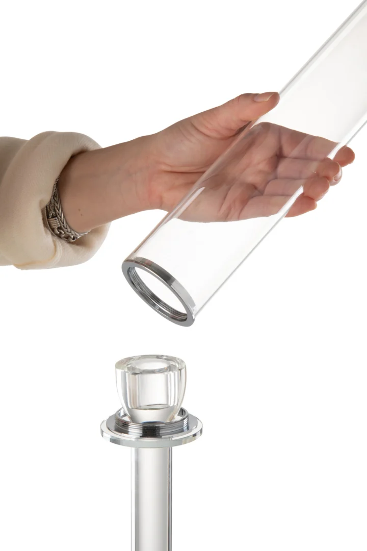 Een hand houdt de Kaarsenhouder Moira Glas Transparant Groot boven een metalen standaard met een glazen stop, klaar om het op de standaard te plaatsen. De achtergrond is wit.