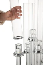 Een hand tilt de Kaarsenhouder 5 Dun Glas Transparant uit een groep vergelijkbare glazen kandelaars op transparante standaards, allemaal tegen een witte achtergrond.