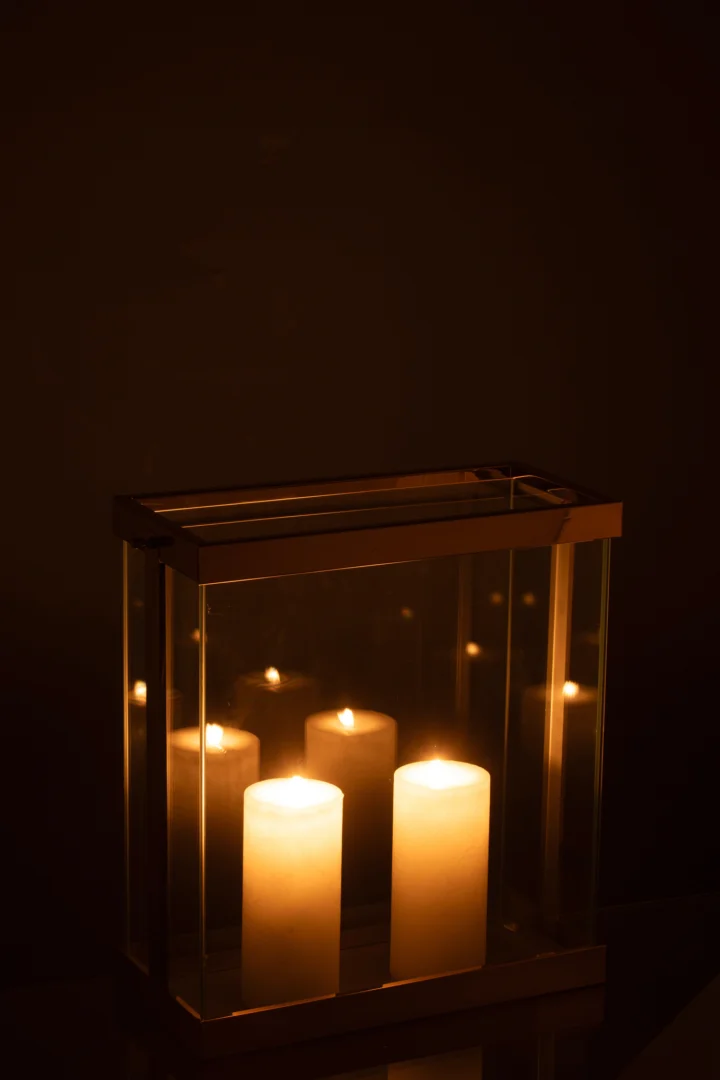 De Kaarsenhouder Lantaarn Rechthoekig Glas Goud Klein heeft vier brandende pilaarkaarsen in een kleine, rechthoekige lantaarn van glas en goud, die een warme, zachte gloed werpt in een donkere omgeving.