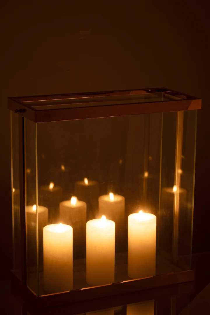 De Kaarsenhouder Lantaarn Rechthoekig Glas Koper Groot houdt vier pilaarkaarsen in een lantaarn van glas en koper, die een warme gloed en zachte reflecties op het glas werpt in een schemerige kamer.