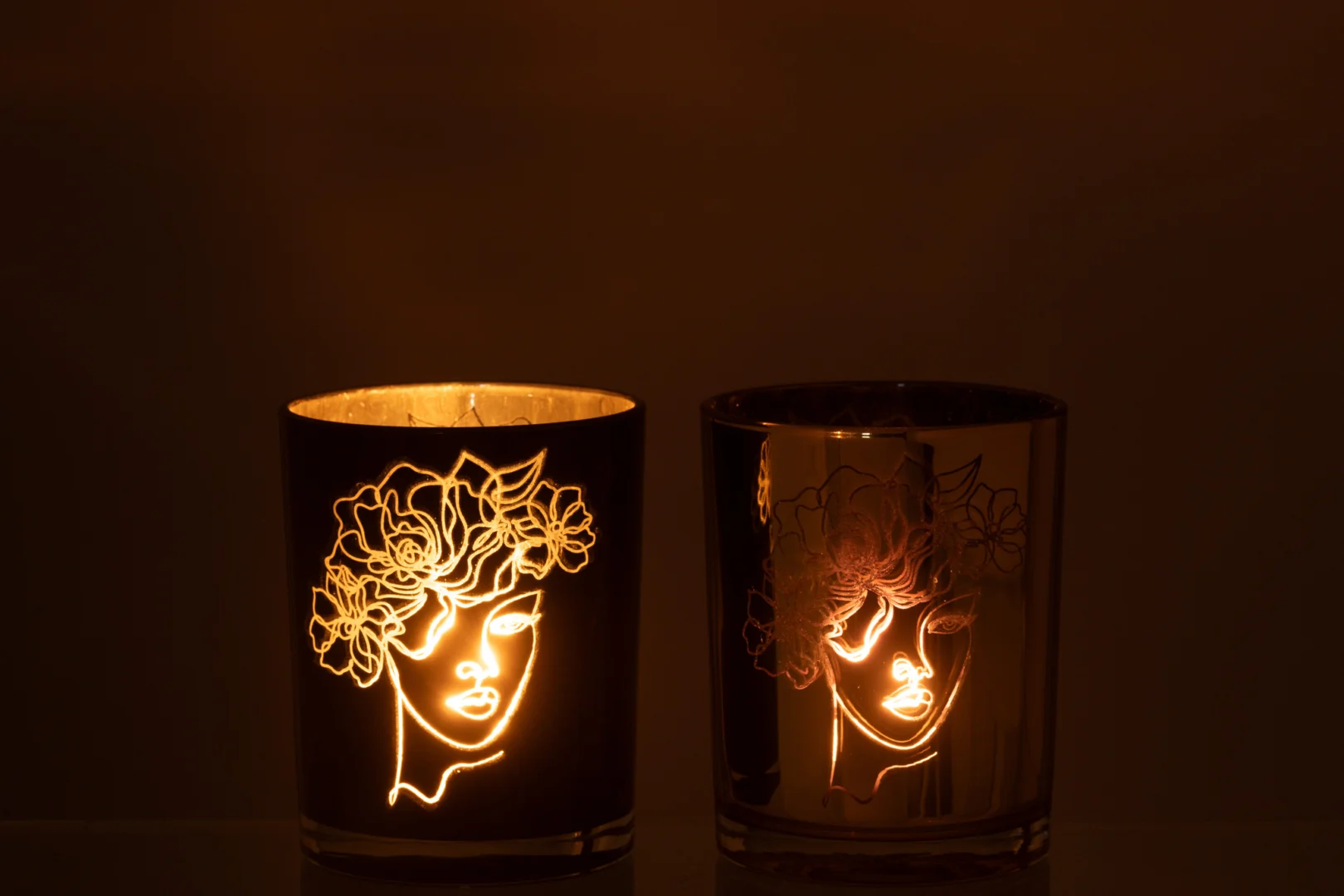 De Kaarsenhouder Orkaan Dame Bloemen Glas Zwart Goud Medium Set 2 heeft twee kaarsen in reflecterende houders met een abstract vrouwengezicht en bloemmotief, waarvan er één brandt om een warme gloed te werpen die het kunstwerk in het donker benadrukt.