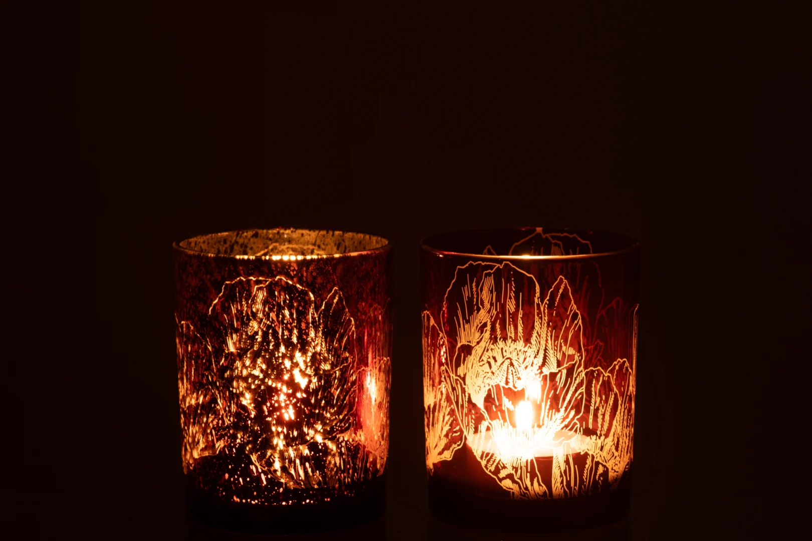 De Kaarsenhouder Hurricane Bloemen Glas Bordeaux Medium Set 2 heeft twee verlichte glazen kandelaars die warm oplichten in het donker en prachtige patronen van licht en schaduw op hun oppervlak werpen.