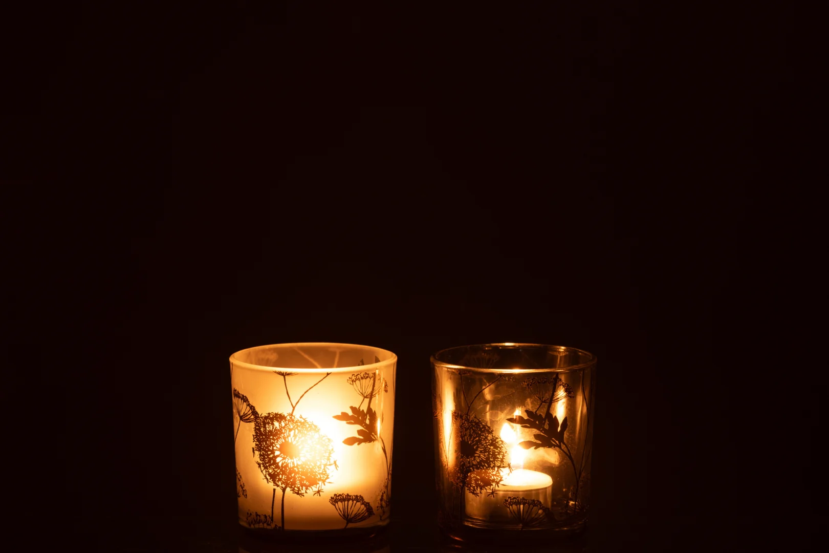 De Theelichthouder Paardenbloem Glas Helder Mat Goud Set 2 werpt een warme, uitnodigende gloed uit de glazen houders met paardenbloemmotieven en creëert een gezellige en rustige sfeer tegen een donkere achtergrond.