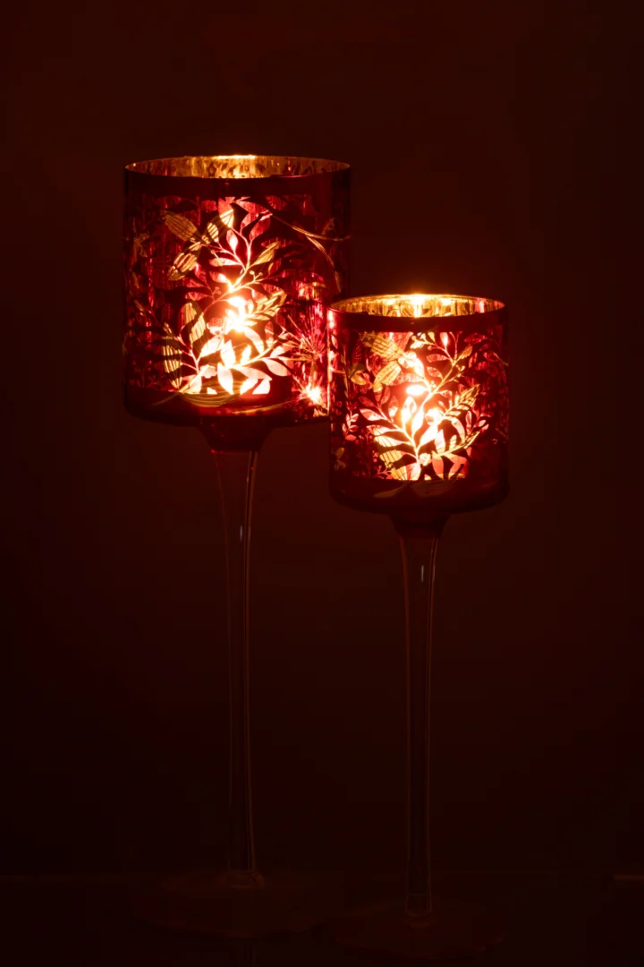 De Theelichthouder Op Voet Bladeren Glas Rood/Antiek Goud Groot heeft roodgetint glas met bladmotieven, verheven op een heldere standaard en met een warme, zachte gloed tegen een donkere achtergrond.