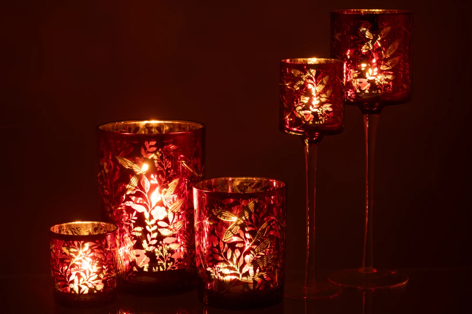 De "Theelichthouder Op Voet Bladeren Glas Rood/Antiek Goud Groot" is een rode glazen kandelaar met antieke bladgoudmotieven, die een warme, decoratieve gloed verspreiden. Twee houders staan op hoge stelen voor een elegante, gezellige sfeer.