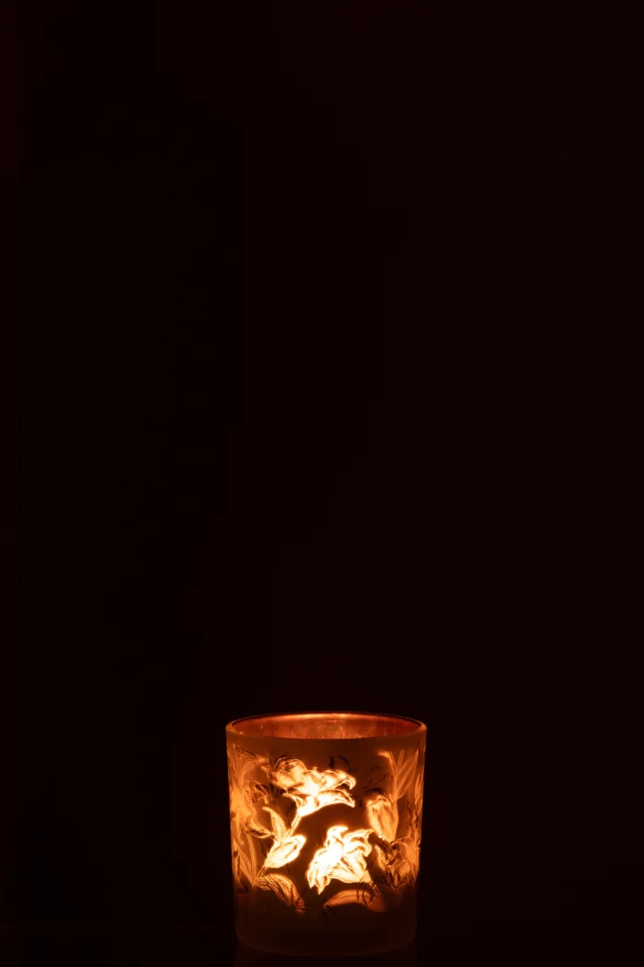 Een enkele Theelichthouder Bloemen Glas Mat Wit/Rood/Goud Klein kandelaar gloeit warm in de duisternis en werpt licht op de bloemmotieven tegen een zwarte achtergrond.