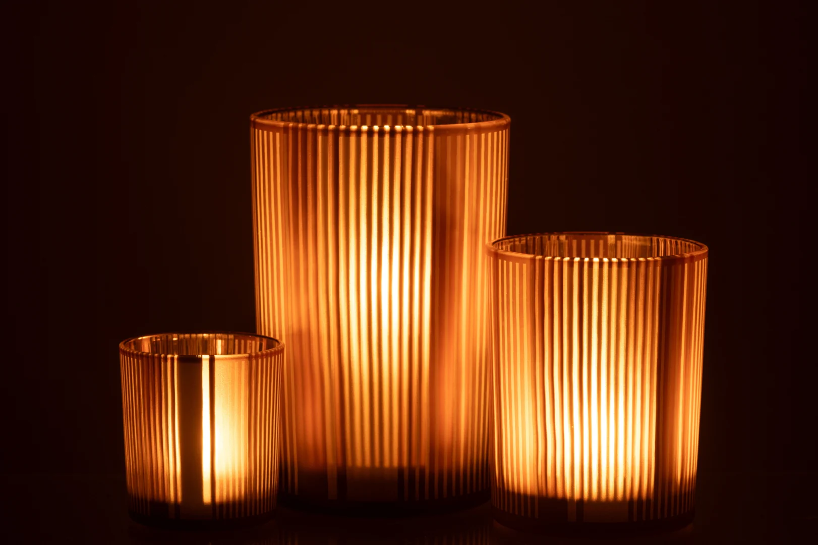 De Kaarsenhouder Orkaan Strepen Glas Bruin Wit Groot straalt warm in een donkere omgeving en werpt zacht gouden licht door het geribbelde glasontwerp en het kenmerkende bruin-wit gestreepte patroon.