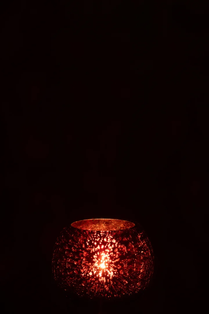 De Theelichthouder Bal Craquelure Glas Roze Klein gloeit warm in de duisternis met een brandende kaars erin en werpt delicate patronen op het roze craquelure glazen oppervlak.