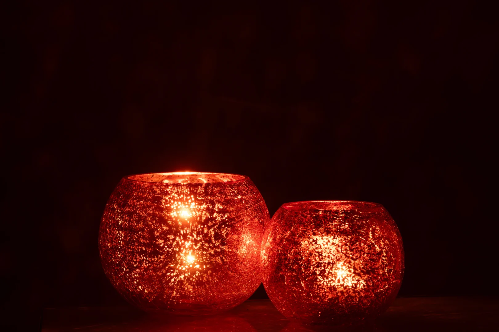 De Theelichthouder Bal Craquelure Glas Oranje Groot straalt een warme oranje gloed uit in het donker, waarbij het ronde, gestructureerde glazen ontwerp een gezellige en uitnodigende sfeer creëert.