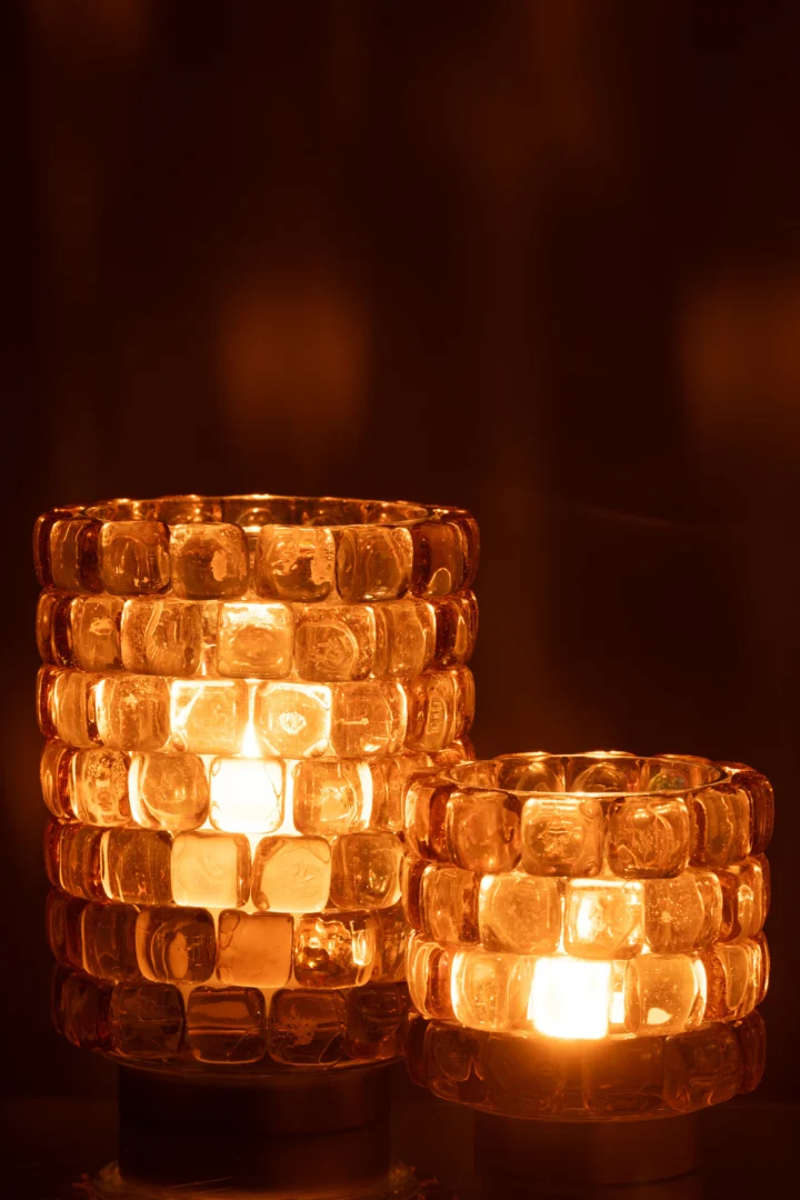 De Kaarsenhouder Hurricane Vierkant Mozaïekglas Cognac Groot gloeit warm met brandende kaarsen, de vierkante mozaïekglasstukjes werpen een zacht amberkleurig licht tegen een donkere, wazige achtergrond.