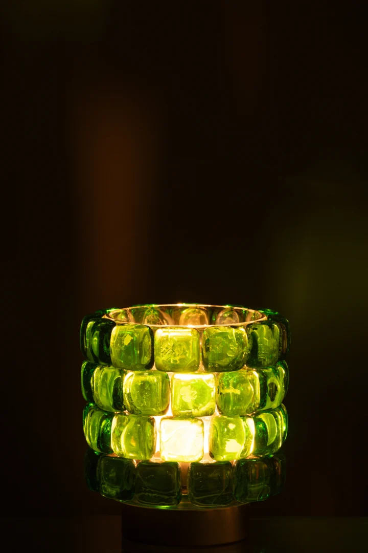 Theelichthouder Vierkant Mozaïekglas Groen Klein, gemaakt van doorschijnende groene glasblokjes en verlicht van binnenuit, verspreidt een warme, sfeervolle gloed tegen een donkere achtergrond.