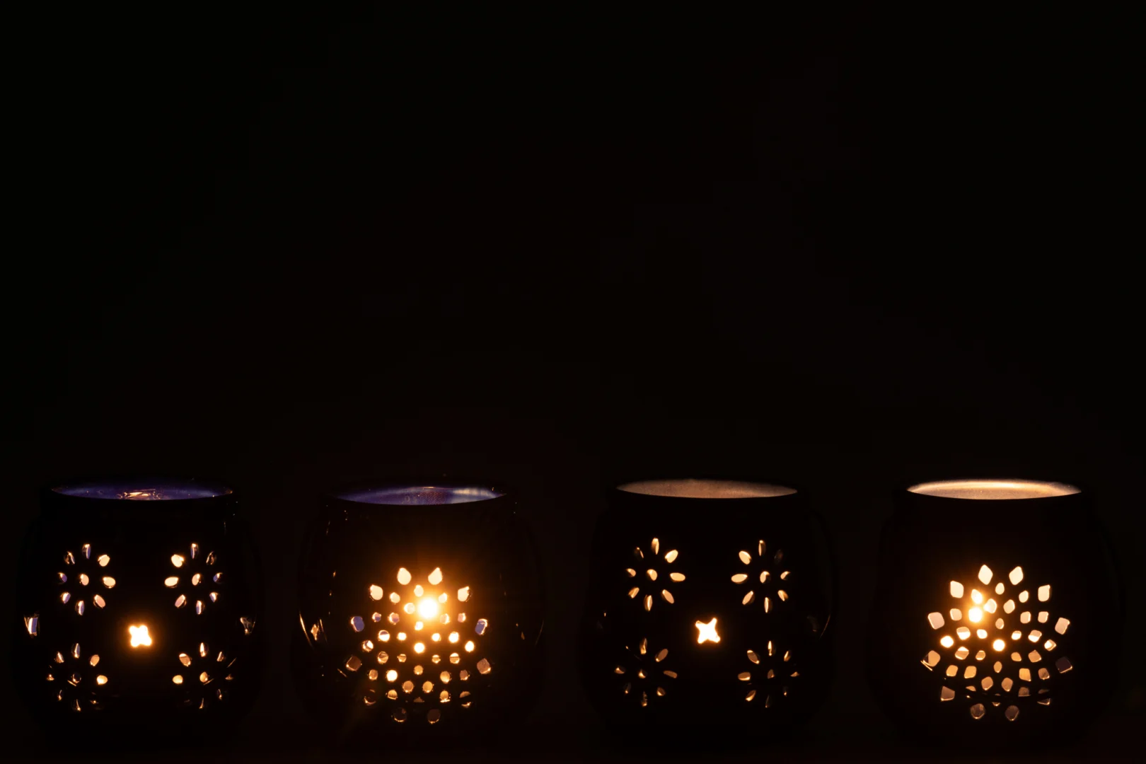 De Theelichthouder Emmer Porselein Blauw Set 4 bestaat uit vier blauwe porseleinen kandelaars met bloemmotieven. Twee stralen een warme gloed uit door de patronen, terwijl twee onverlicht blijven, allemaal op een rij tegen een zwarte achtergrond.