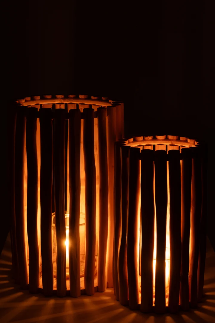 De Kaarsenhouder Lantaarn Teak Natuurlijk Set 2 bestaat uit twee teakhouten lantaarns met verticale lamellen die naast elkaar staan en warm oplichten in het donker met brandende kaarsen erin, waardoor ze een zacht licht en zachte schaduwen werpen.