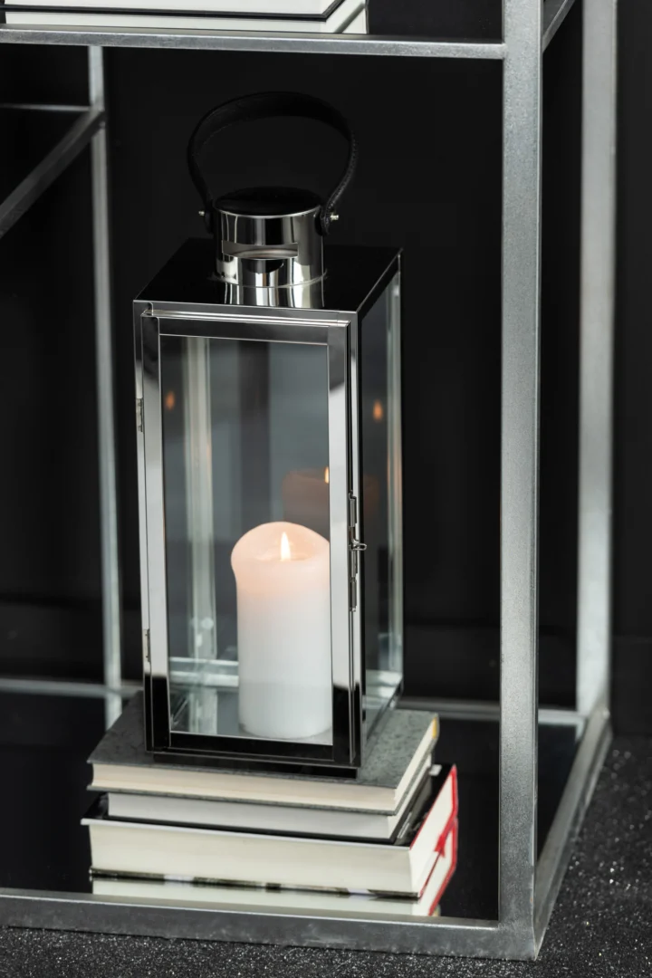 Een Lantaarn Vierkant Metaal Glas Zilver met een brandende witte kaars zit op gesloten boeken op de onderste plank van een moderne metalen stelling tegen een donkere achtergrond.