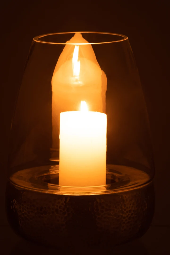 De Kaarsenhouder Orkaan Aardewerk Glas Zilver houdt een gloeiende kaars vast, die een warm, zacht licht werpt en de reflectie ervan op het glazen oppervlak in het donker verlicht.