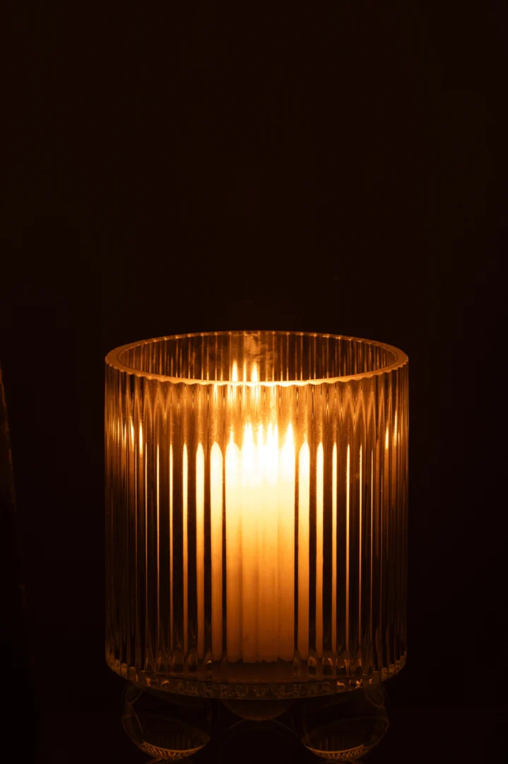 De Kaarsenhouder Orkaan Clara Glas Klein, met zijn heldere geribbelde glas, gloeit warm in het donker wanneer hij wordt aangestoken door een witte kaars en werpt zacht licht en zachte schaduwen om zich heen.