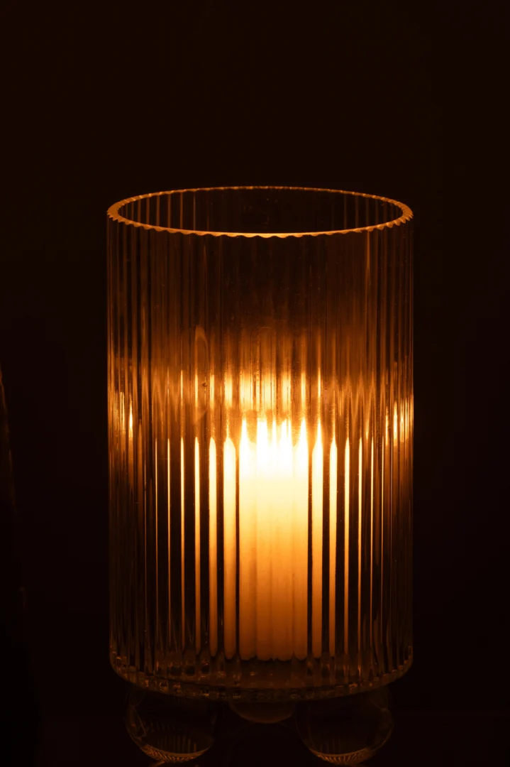 De Orkaan Clara Glas Transparant Groot - Kaarsenhouder heeft een brandende kaars die warm gloeit in het hoge, geribbelde glazen ontwerp, waardoor een gezellige en sfeervolle sfeer wordt gecreëerd tegen een donkere achtergrond.
