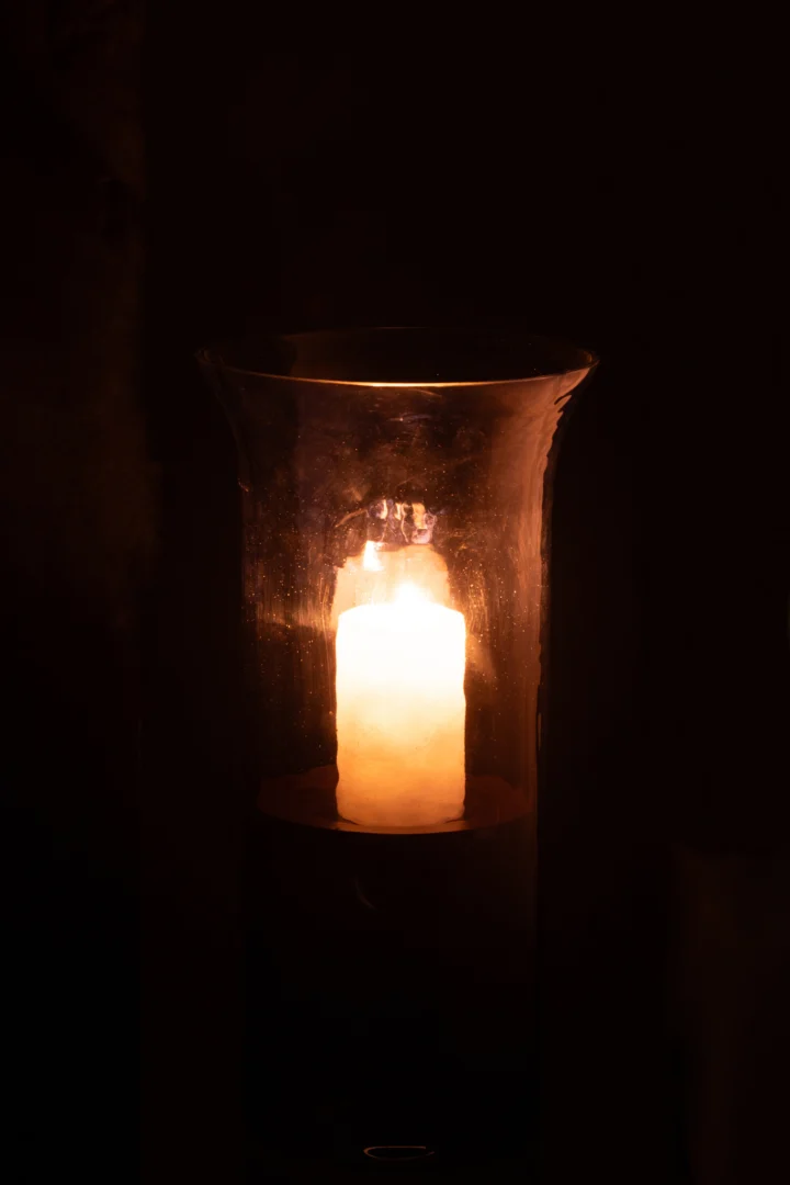 Een brandende kaars gloeit warm in de Orkaan Tarifa Smal Glas Blauw Medium - Kaarsenhouder, de blauwe glazen houder verlicht zachtjes de donkere omgeving.