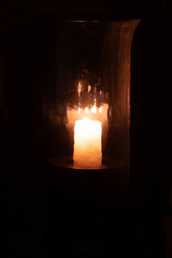 De Orkaan Tarifa Smal Glas Blauw Extra Groot - Kaarsenhouder houdt een brandende kaars vast en werpt een warme, zachte gloed door het blauwe glas in een anders donkere ruimte.