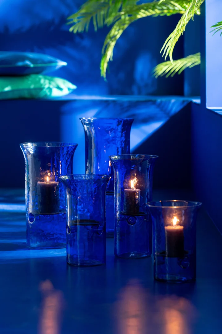 Vijf Orkaan Tarifa Smal Glas Blauw Extra Groot kandelaars met brandende kaarsen staan op een blauw vlak en stralen een warme gloed uit. Groene bladeren en zacht blauw licht versterken de rustige, moderne sfeer op de achtergrond.