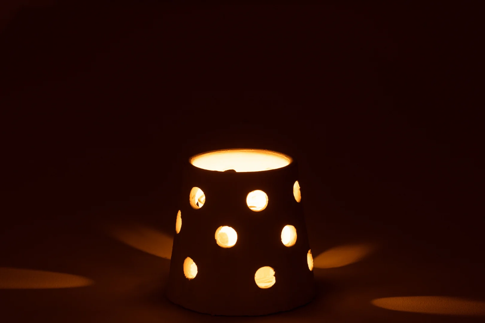 Theelichthouder Ibiza Klei/Glas Wit Smal: een brandende kaars in een witte houder met ronde uitsparingen, die warme lichtpatronen werpt tegen een donkere achtergrond.
