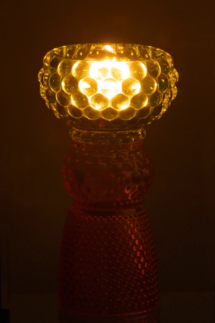 De Kaarsenhouder Jazzy Glas Mix heeft een brandende kaars in een ronde, bubbelende glazen houder op een hoge, gedessineerde rode glazen voet, prachtig weergegeven tegen een donkere achtergrond.