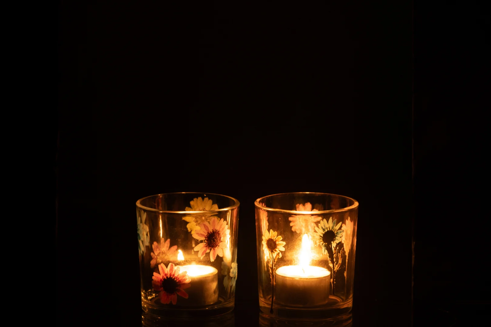 De Kaarsenhouder Posy Glazen Mix Set van 2 heeft twee glazen kandelaars versierd met kleurrijke bloemen, die een warme, gezellige gloed werpen en een vredige sfeer creëren tegen een donkere achtergrond.