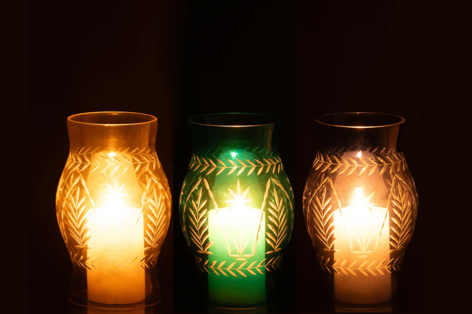 De Orkaan Tarifa Glas Mix Set van 3 - Kaarsenhouders heeft drie decoratieve glazen houders met geëtste bladmotieven, elk oplichtend in gele, groene en amberkleurige tinten tegen een donkere achtergrond.