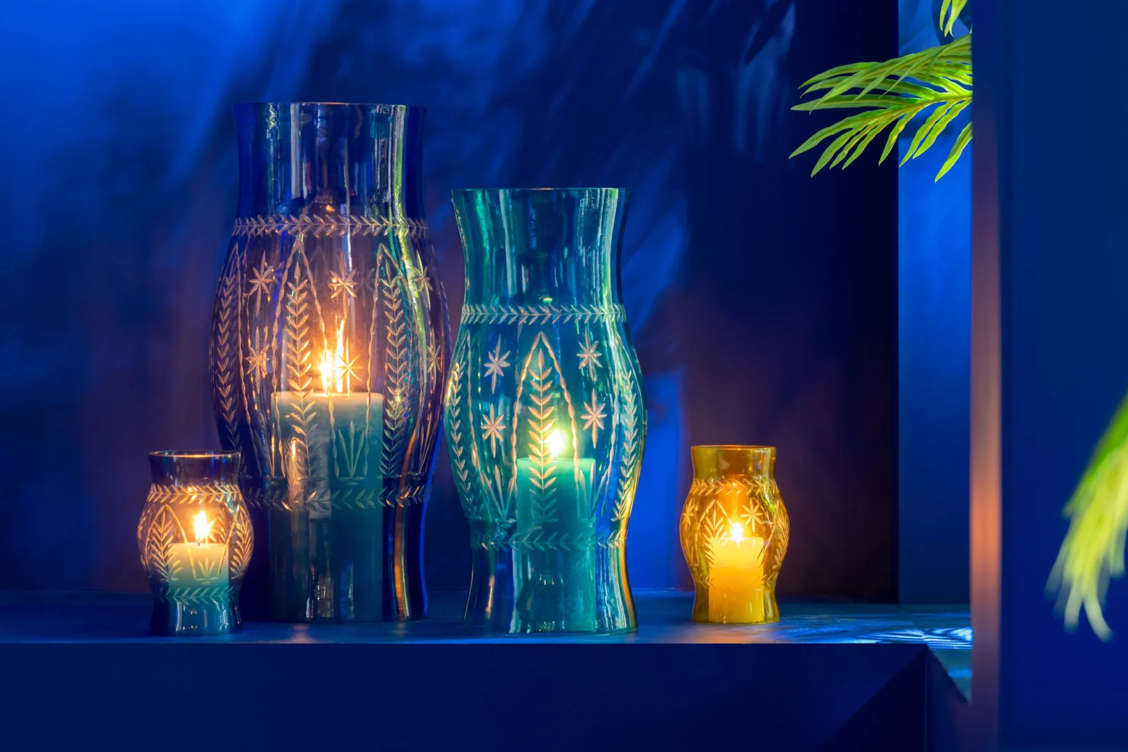 De Orkaan Tarifa Glas Mix Set van 3 kaarsenhouders in blauw, groen en geel schittert met brandende kaarsen tegen een levendige blauwe achtergrond met een groene plant op de achtergrond.