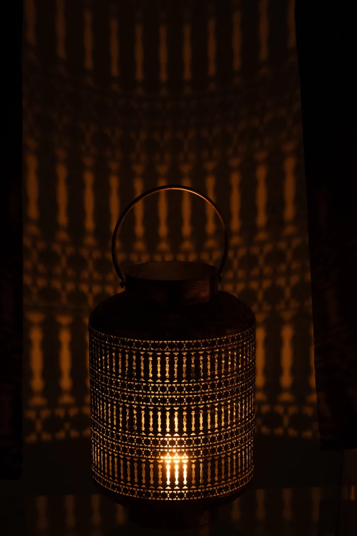 De Lantaarn Cilinder Antiek IJzer Brons Klein heeft een handvat en straalt warm licht uit, waardoor ingewikkelde schaduwen met patronen op de donkere muur erachter worden geworpen.