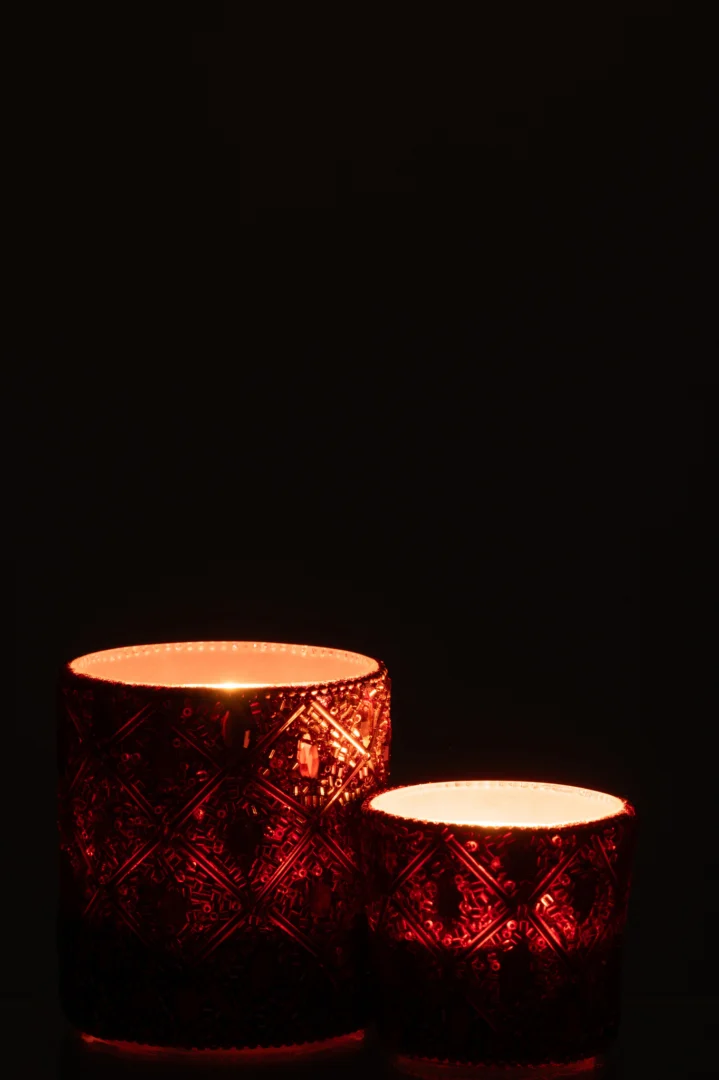 Een grote, donkerbruine glazen kandelaar met kralen gloeit warm in het donker en werpt zacht licht tegen een zwarte achtergrond. Dit is de Theelichthouder Glas Kralen Donkerbruin Roestbruin Groot, links geplaatst.