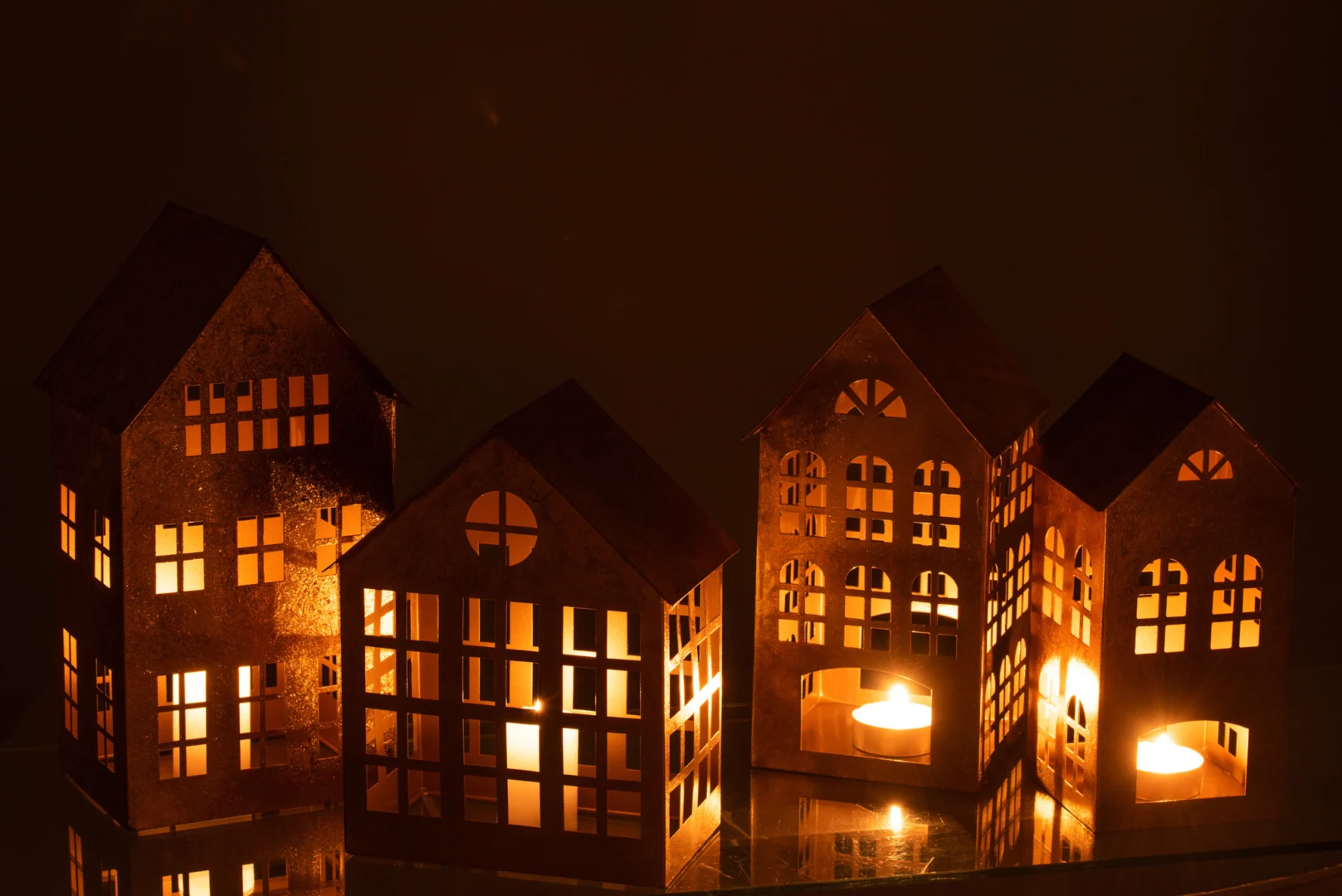 Vier lantaarns van Kaarsenhouder Huis Sigma IJzer Goud, in de vorm van kleine huisjes en gemaakt van goudkleurig ijzer, staan op een rij met kaarsen erin en werpen een warme gloed en zachte schaduwen in een donkere kamer.