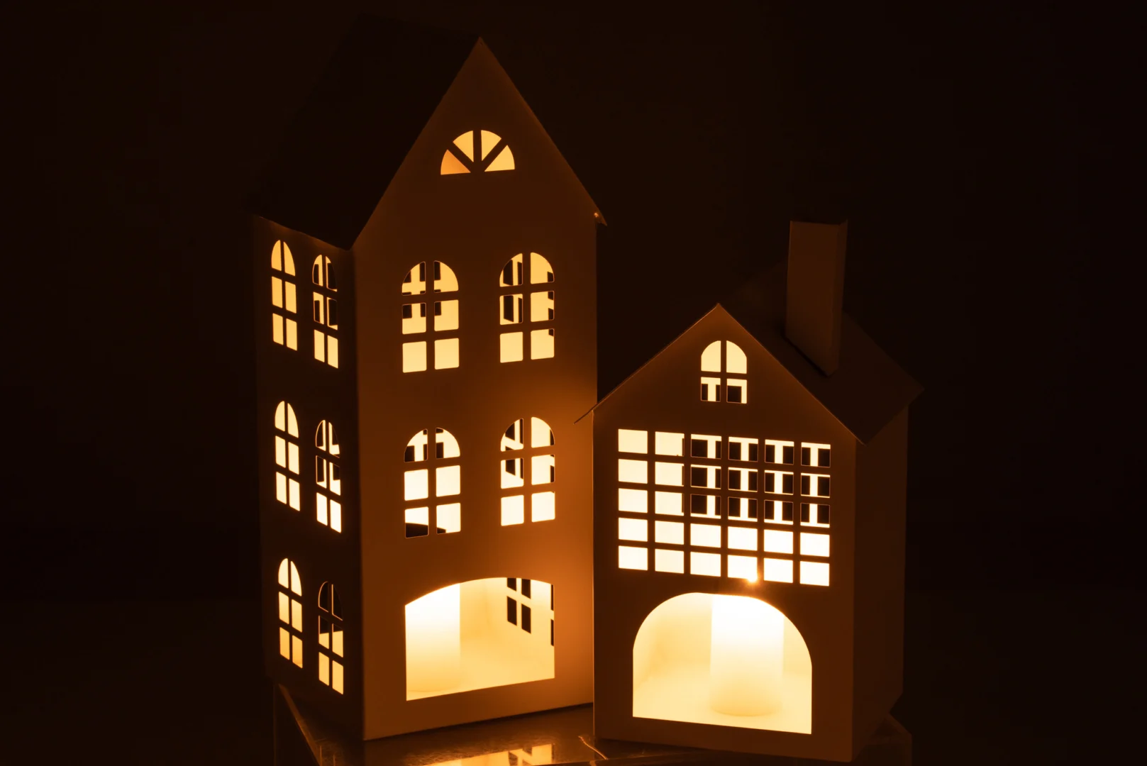 Het Kaarsenhouder Huis Psi IJzer Wit is een decoratief wit ijzeren huis dat van binnenuit warm gloeit en zachtgeel licht werpt door de ramen en deuren tegen een donkere achtergrond.