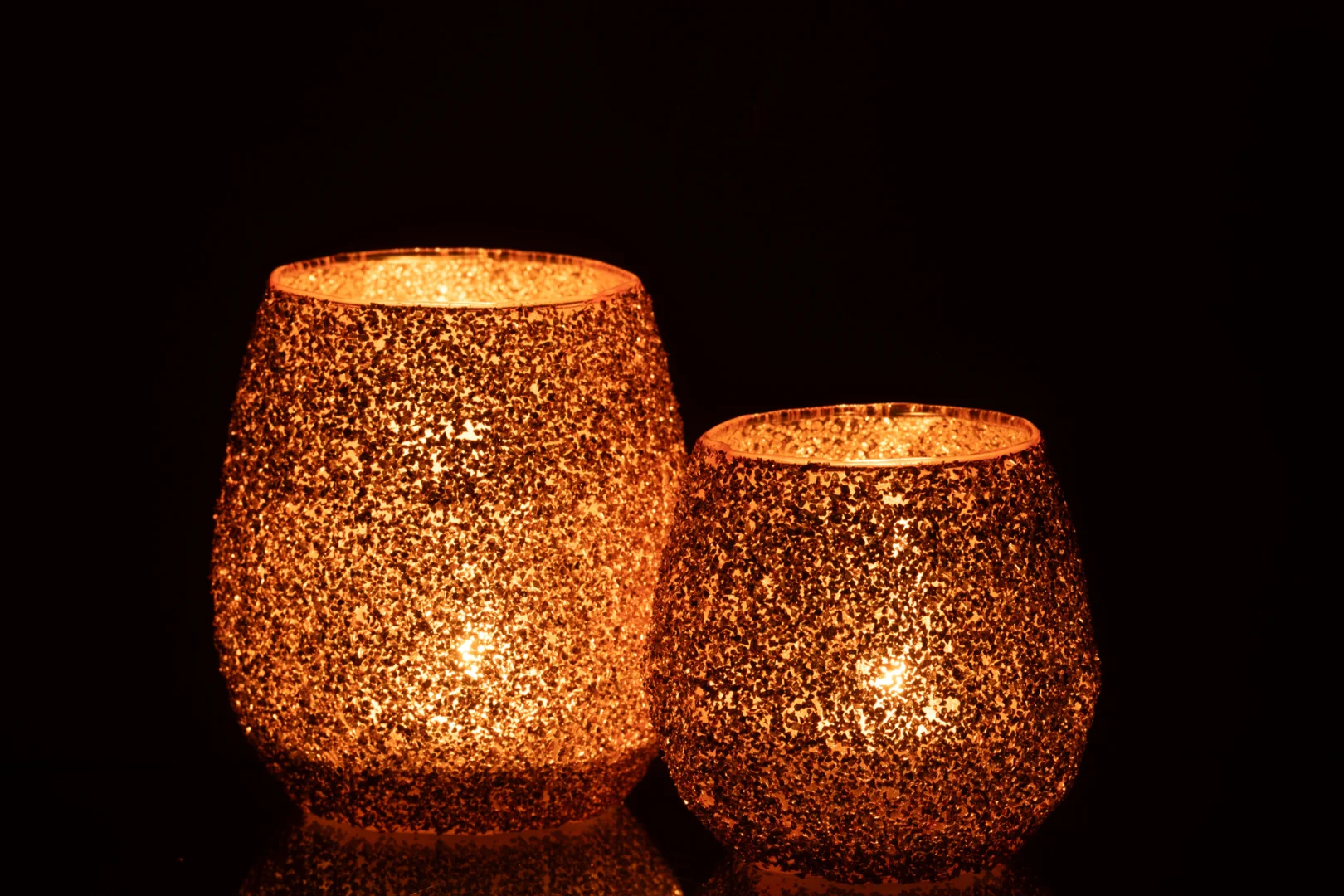 De Theelichthouder Glas Perzik Groot kandelaars gloeien warm op met brandende kaarsen erin, hun gestructureerde glazen oppervlakken fonkelen goud en werpen een zacht, gezellig licht tegen een donkere achtergrond.