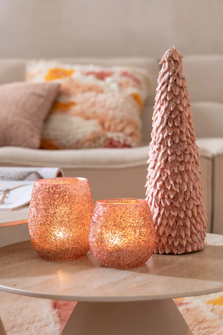 Twee Theelichthouder Glas Perzik Groot kandelaars en een hoge roze decoratieve boom met structuur staan op een ronde houten salontafel in een gezellige woonkamer met een crèmekleurige bank en kleurrijke kussens op de achtergrond.