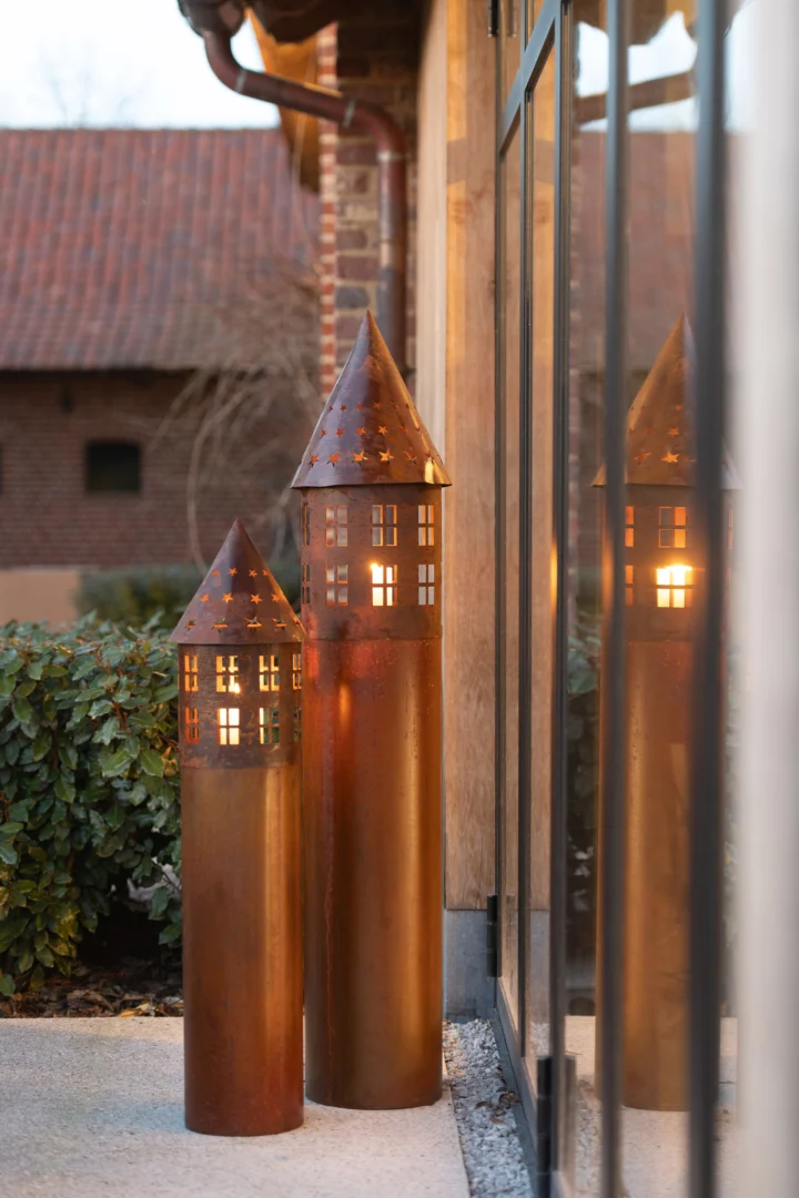 Twee hoge lantaarns van Kaarsenhouder Ster IJzer Roestbruin met puntdaken en uitgesneden sterramen stralen warm naast een glazen buitenmuur, met op de achtergrond groen en bakstenen gebouwen.