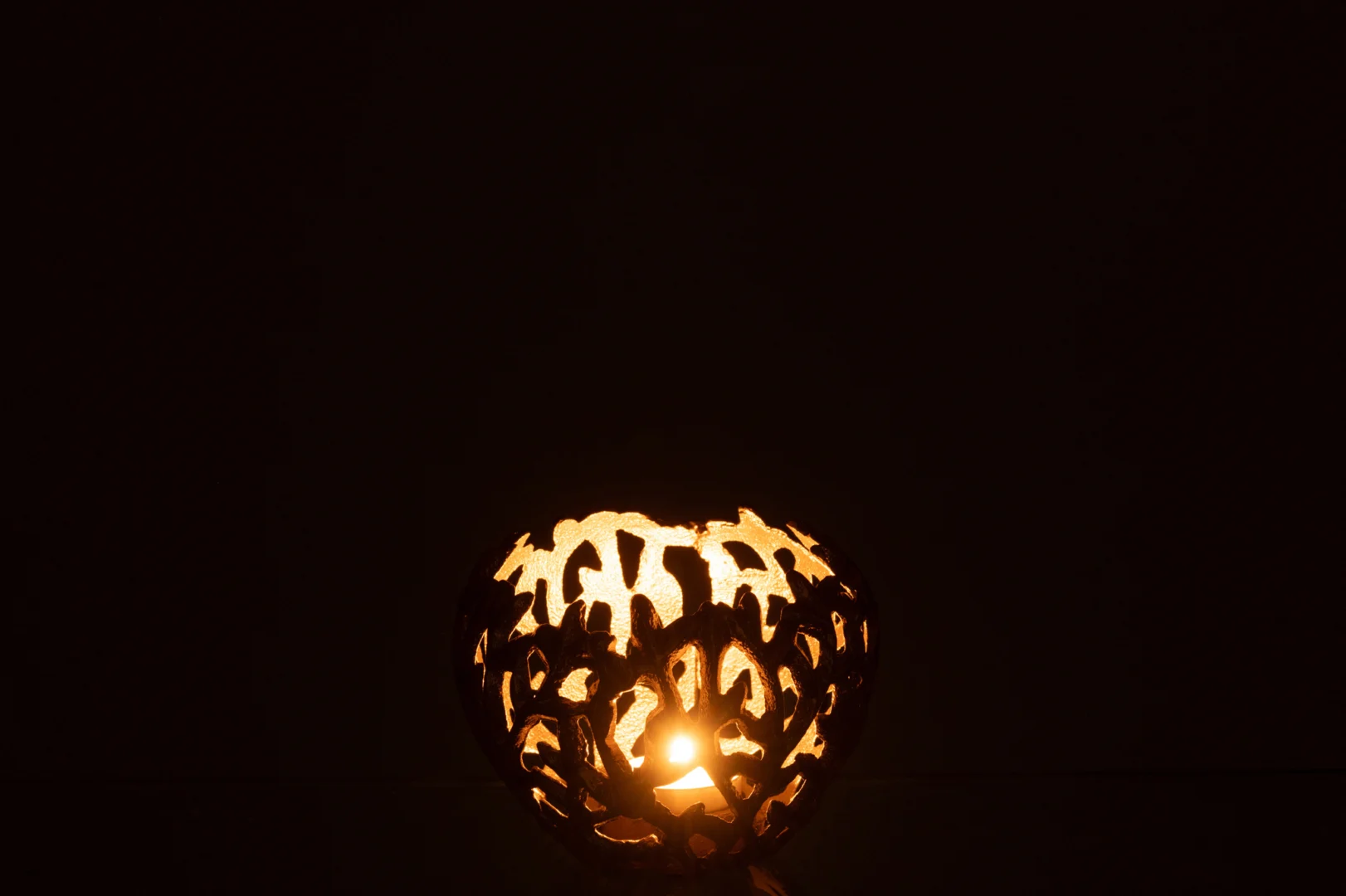 Een brandende kaars in de Kaarsenhouder Blad Aluminium Goudkleurig Klein werpt ingewikkelde schaduwen en patronen op een donkere achtergrond.