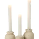 Drie witte pilaarkaarsen branden in de Kaarsenhouder Cement Lichtbeige Set van 3 gestructureerde, lichtbeige keramiek houders van verschillende hoogtes tegen een effen witte achtergrond.
