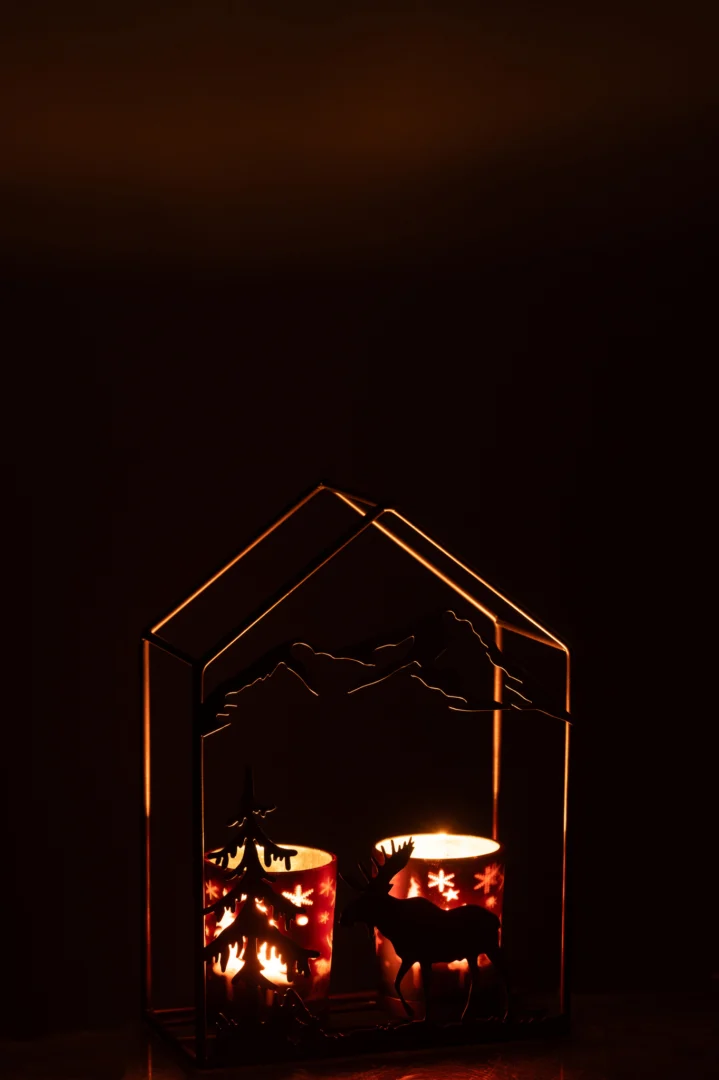 De Theelichthouder Huis IJzer Roestbruin/Bruin 2 Glazen heeft twee brandende kaarsen in houders met stervormig patroon in een huisvormig metalen frame, dat een warme gloed werpt met berg-, boom- en hertensilhouetten in het donker.